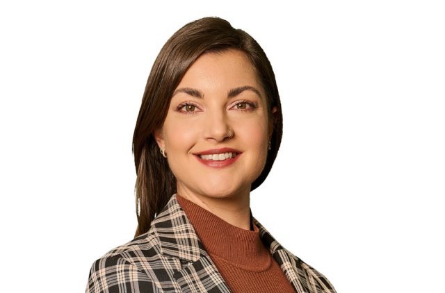 Alt text: A professional portrait of a woman standing against a white background. She has straight dark brown hair and is smiling at the camera. She is wearing a black and beige plaid blazer over a rust-colored turtleneck sweater. The image appears to be a formal or business profile photo, likely for a company or corporate setting.