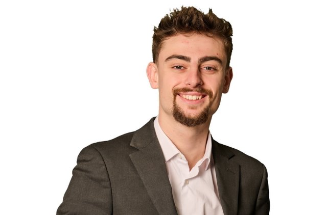 A professional portrait of a young man standing against a white background. He is smiling and wearing a dark grey suit with a white dress shirt. His left hand is in his pocket and he has short, styled hair and a trimmed beard. The image is likely intended for business or corporate use.