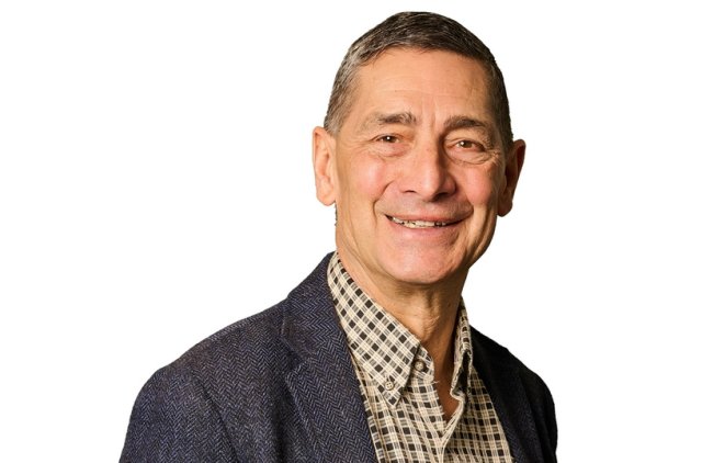 A professional portrait of a smiling older man standing against a white background. He is wearing a dark blue textured blazer over a beige and black plaid button-down shirt. His posture is relaxed and confident, making the image suitable for a business or corporate profile.