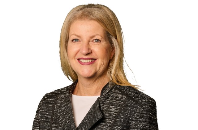 A professional portrait of a smiling woman standing against a white background. She has shoulder-length blonde hair and is wearing a textured, dark gray and black jacket over a white top. Her hands are gently clasped in front of her. The image appears to be taken for a formal or business-related profile.