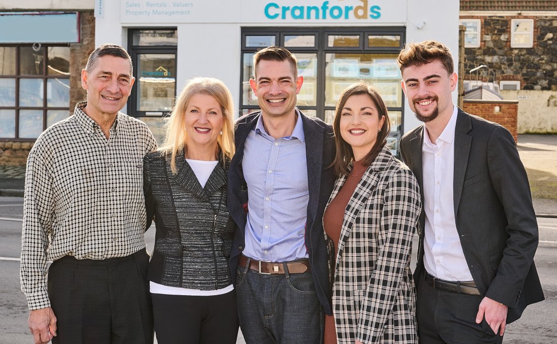 A group of five people standing closely together and smiling outside a building with a sign that reads "Cranfords" and "Estate Agents" offering services like sales, rentals, valuations, and property management. The building has a white facade with large windows and a bay window on the top floor. The group appears to be dressed in business casual and formal attire, suggesting they may be staff or associates of the estate agency.
